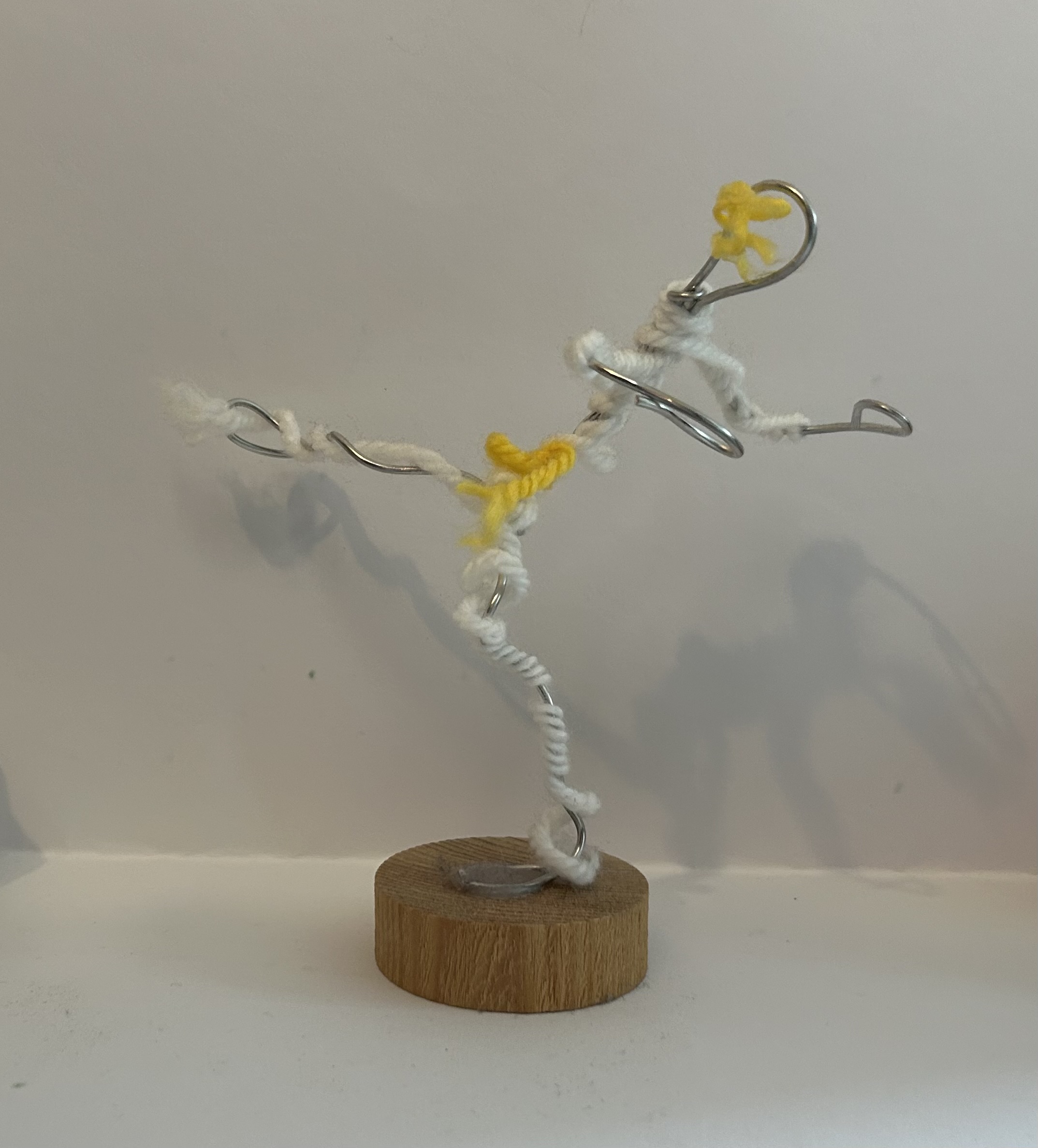 a figure on a round wood block. the figure is made of wire with white and yellow string around it. the figure looks like a kung fu fighter kicking. made by a child.